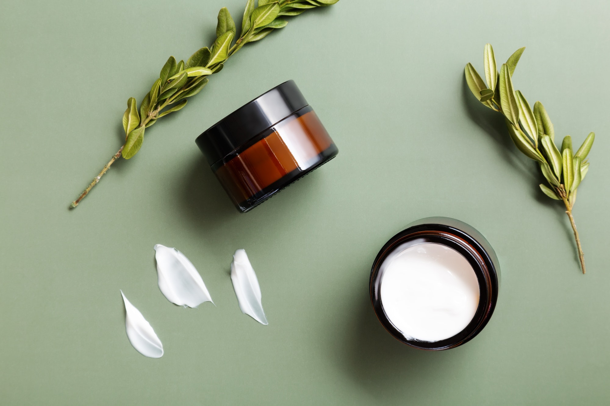 Glass jars of moisturizing cream and plants on green background. Concept of natural cosmetics