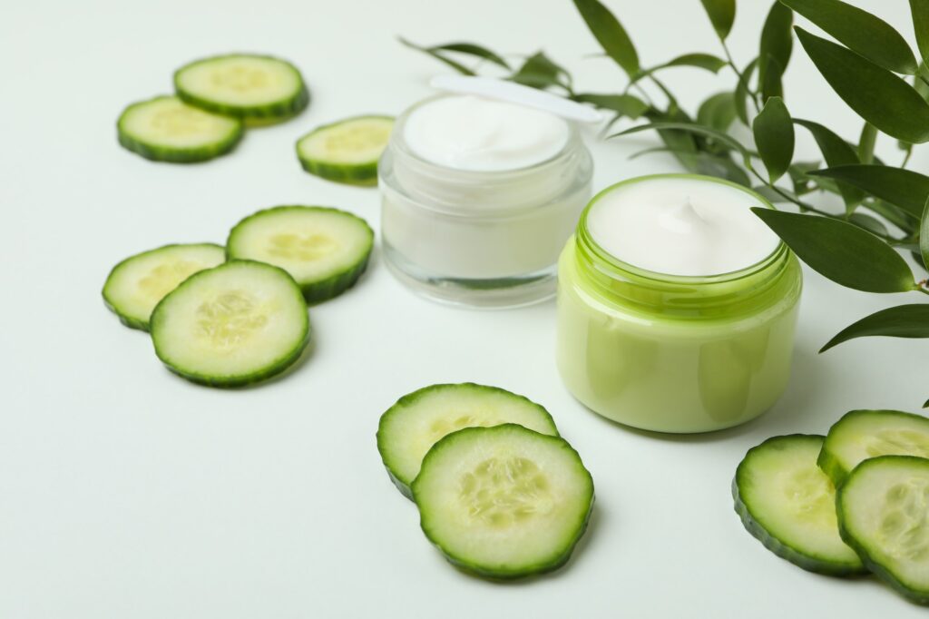 Jars with cosmetic cream, cucumber slices and branch on white table